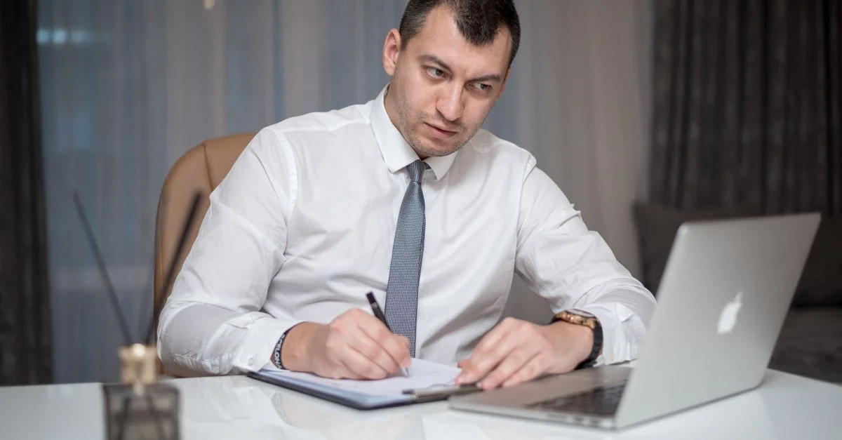 Homme d'affaires concentré en chemise blanche prenant des notes à son bureau avec un ordinateur portable.