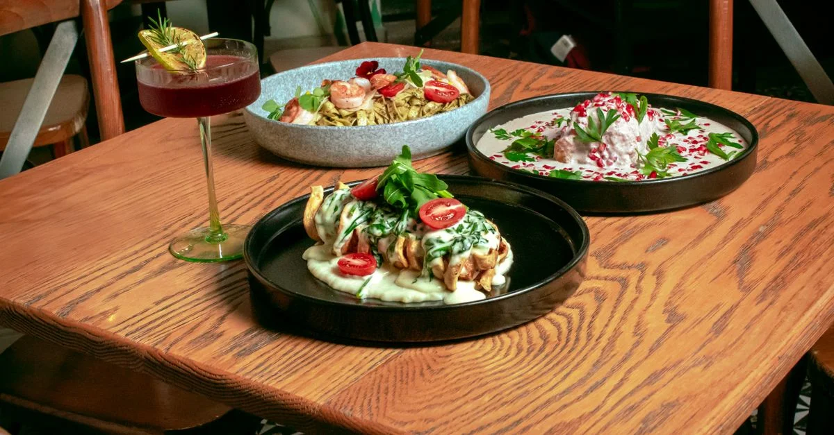 Trois plats gastronomiques avec un cocktail servis sur une table en bois, mettant en valeur l'art culinaire dans un cadre de restaurant cosy.