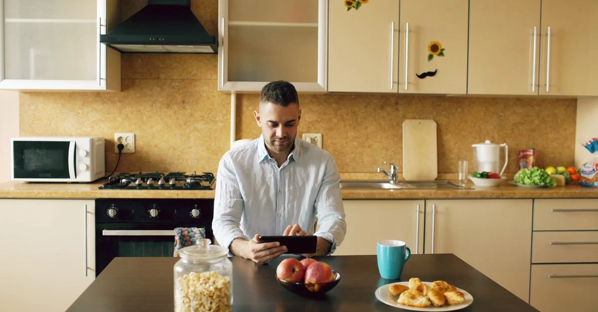 Homme assis à table dans la cuisine utilisant une tablette, entouré d'articles de petit-déjeuner et d'appareils de cuisine modernes.
